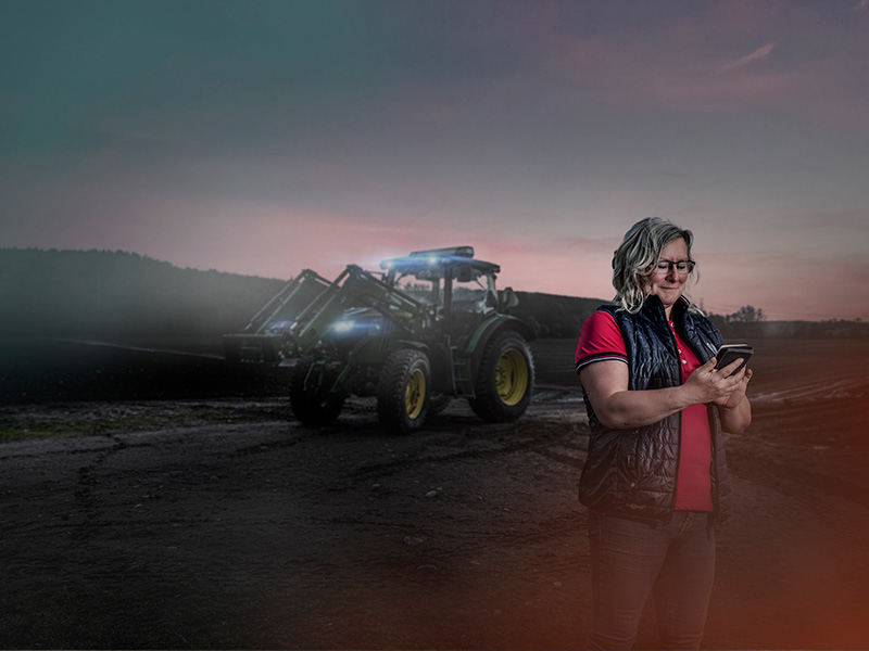 Kvinna läser mobil på en åker med traktor i bakgrunden, skymning